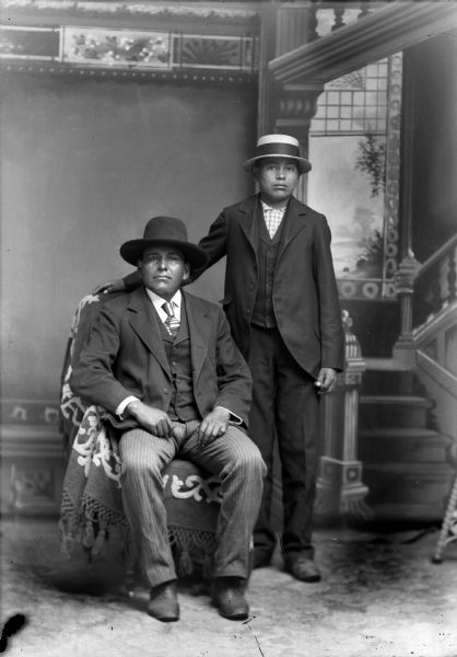 Studio portrait in front of a painted backdrop of a Ho-Chunk man posing siting on the left wearing a suit, necktie, and hat, and a younger Ho-Chunk man posing standing on the right wearing a suit, plaid shirt, and straw hat, and with his right arm resting on the back of the chair.