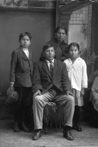 Full-length studio portrait of a Ho-Chunk family. In the background is a painted backdrop. Names (sitting) David Bow Littlesoldier (Maw Jhay Mau Nee Kah). Standing (l to r) Simon Robert Littlesoldier (Wo No Ka Re Hun Kah) (Son of David Bow Littlesoldier [Maw Jhay Mau Nee Kah] and Emma Thunder, Littlesoldier [We Pa Ma Ka Ra Win Kah]), Emma Thunder, Littlesoldier (We Pa Ma Ka Ra Win Kah) and Ida Ada Littlesoldier, Whitedog (Ka Ro Cho Win Kah).