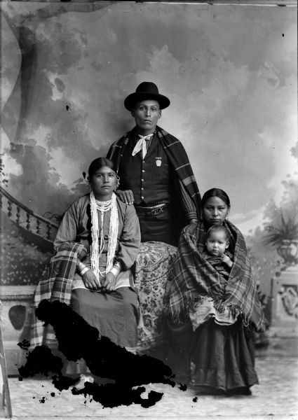 Studio portrait in front of a painted backdrop of two Ho-Chunk women posing sitting, one holding an infant in her lap, and a man standing. The woman on the left is sitting on a prop stone wall and is wearing several necklaces, earrings, and file bracelets, and holding a shawl draped over her left arm, while the woman on the right is wearing several earrings and is wrapped in a shawl and holding a Ho-Chunk child. The Ho-Chunk man posing standing has his right hand on the left shoulder of the woman on the left, and is wearing a vest, bandana, hat, and a plaid shawl over his shoulders.