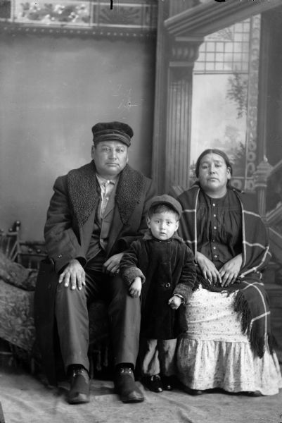 Studio portrait in front of a painted backdrop of a small Ho-Chunk boy posing standing and wearing a long winter coat and hat. The boy is between a Ho-Chunk man posing sitting on the left, who is wearing a long winter coat and hat, and a Ho-Chunk woman posing sitting on the right, who is wearing a light-colored skirt and dark-colored blouse with a plaid fringed shawl over her shoulders.