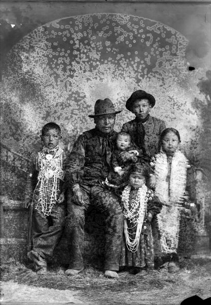 Studio portrait in front of a painted backdrop of a Ho-Chunk man posing sitting wearing a vest, bandana, and hat, and holding an infant. He is surrounded by Ho-Chunk children. A boy is posing standing on the left wearing bandoleers, a gorget, and several necklaces. On the right a Ho-Chunk boy is posing standing and wearing a suit, bandana, and hat, and in front of him a small Ho-Chunk girl is posing standing with several necklaces. On the right is a young Ho-Chunk woman posing and wearing several long necklaces.