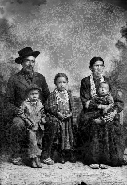 Studio portrait of a family posing in front of a painted backdrop. On the left is a Ho-Chunk, or mixed blood (African or European) man sitting on the left wearing a suit, bandana, moccasins, and hat. He is holding the arm of a small Ho-Chunk child standing in front of him, who is wearing a suit and hat. A Ho-Chunk girl is standing in the center, and is wearing several necklaces and a shawl over her shoulders. On the right is a Ho-Chunk woman is sitting and wearing several necklaces, earrings, moccasins, and file bracelets, and holding a small child on her lap.