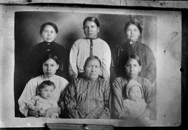 Copy photograph of a studio portrait of three Ho-Chunk women posing sitting. The women on the left and right are holding small children on their laps. Behind them three Ho-Chunk women are posing standing. All the are wearing contemporary clothing.
