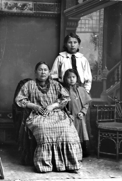 Studio portrait in front of a painted backdrop of a Ho-Chunk woman posing sitting on the left. She is wearing several necklaces, earrings, and a plaid dress. Behind her on the right a young Ho-Chunk woman posing standing and wearing a light-colored blouse. In front on the right is a Ho-Chunk girl posing standing and wearing a dark winter coat.