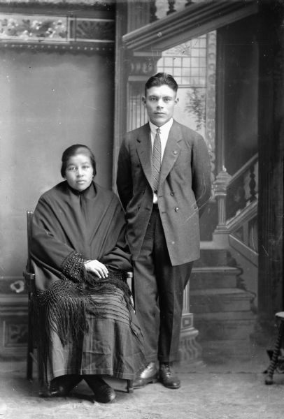 Studio portrait in front of a painted backdrop of a Ho-Chunk woman wrapped in a fringed shawl posing sitting on the left, and a Ho-Chunk man posing standing on the right and wearing a suit and necktie.