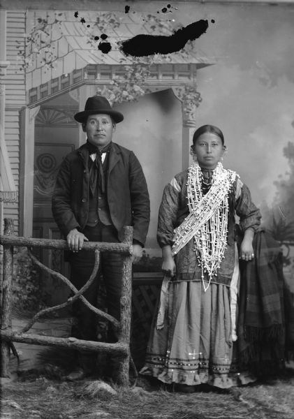 Studio portrait in front of a painted backdrop of Tom Whitewater, standing behind a prop wooden fence, and his wife, Sallie Dora Goodvillage, Whitewater (Wa Nik Shootch Win Kah) (Red Bird Woman). Tom is wearing a suit, bandana, and hat. Sallie is wearing several necklaces, more necklaces as a bandoleer, earrings, and rings.