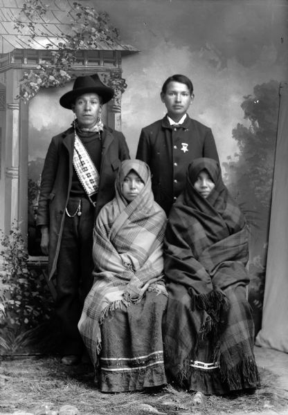 Studio portrait in front of a painted backdrop of two Ho-Chunk women posing sitting and wrapped in fringed plaid shawls which only show their faces. Behind them two Ho-Chunk men are posing standing. The man on the left is wearing a bandoleer, earrings, and hat, and the man on the right is wearing a winter coat and star insignia.