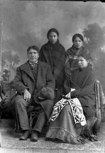 Studio portrait in front of a painted backdrop of a Ho-Chunk man posing sitting on the left and wearing a suit, necktie, winter overcoat, and holding a hat in his right hand. On the right is a Ho-Chunk woman posing sitting and wrapped in a ribbon work shawl. Standing behind them are two young Ho-Chunk women wrapped in plaid shawls.