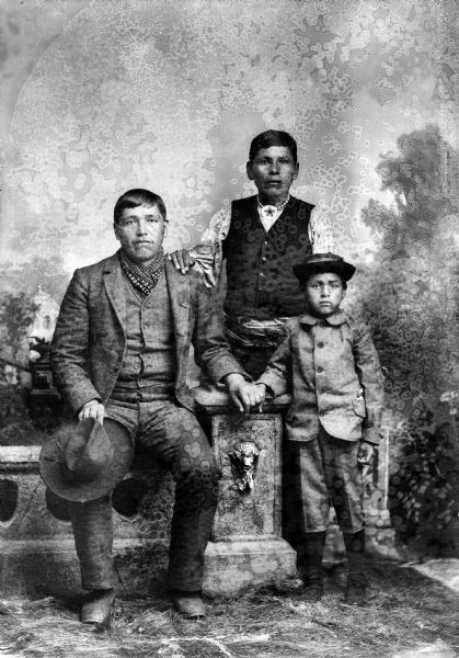 Studio portrait in front of a painted backdrop of a Ho-Chunk man posing sitting on a prop stone wall on the left, and wearing a suit and bandana and holding a hat in his right hand. He is holding the hand of a boy posing standing on the right who is wearing a light-colored suit and a hat. A man is standing behind them wearing a dark-colored vest and a gorget. He has his right hand on the other man's shoulder.