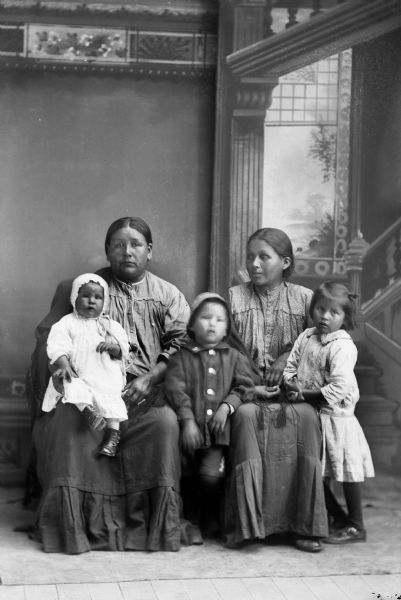Studio portrait in front of a painted backdrop of two Ho-Chunk women posing sitting and wearing contemporary dresses. The woman on the left is holding a small Ho-Chunk child on her right leg, and two other Ho-Chunk children are posing, one standing in the center between the two women, and the other standing on the far right.