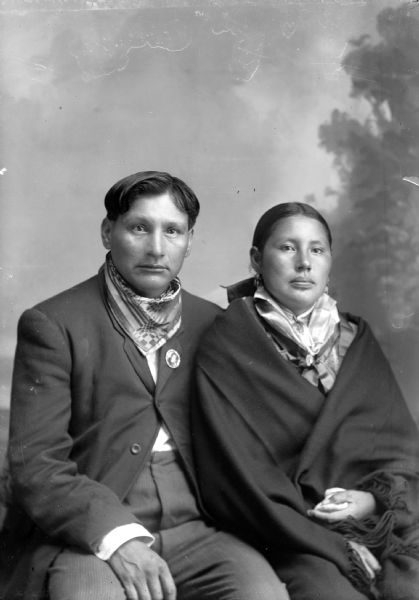 Three-quarter length studio portrait in front of a painted backdrop of a Ho-Chunk man with short hair posing sitting on the left wearing a suit coat and bandana, and a Ho-Chunk woman sitting on the right wrapped in a fringed shawl, and wearing a bandana, and earrings.