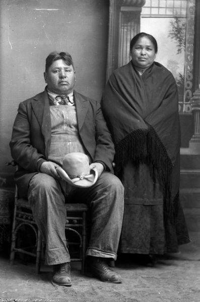 Studio portrait in front of a painted backdrop of a Ho-Chunk man posing sitting on the left, wearing bib overalls, a suit coat, and holding a hat in both hands. On the right is a Ho-Chunk woman posing standing, wrapped in a fringed shawl and wearing a dark-colored dress.
