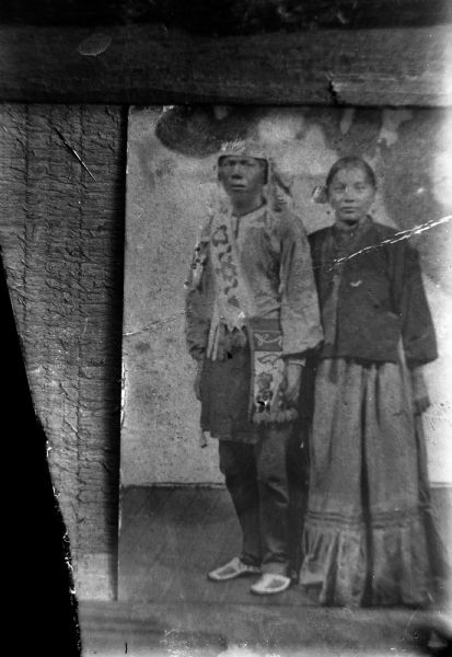 Copy photograph of a studio portrait of a Ho-Chunk man posing standing on the left wearing a floral pattern bandoleer, turban, and Sioux moccasins, and a Ho-Chunk woman posing standing on the right wearing a dark-colored blouse, light-colored skirt, and several necklaces.