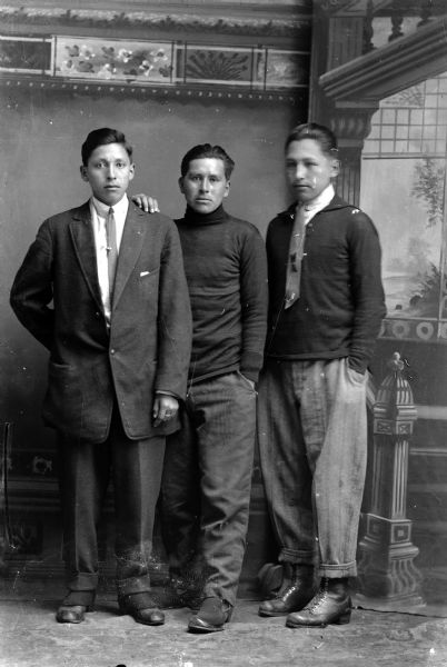 Studio full-length portrait in front of a painted backdrop of three Ho-Chunk men posing standing. The man on the left is wearing a suit and necktie, the man in the center is wearing a turtleneck sweater, and the man on the right is wearing a sweater and thin scarf.