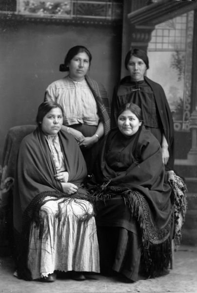 Studio portrait in front of a painted backdrop of two Ho-Chunk women  sitting wrapped in fringed shawls, in front of two Ho-Chunk women standing with shawls over their left shoulders. They are identified as Lucy Thunderking Lowe Yellowbank (MaHiskaWinKah), standing left, and Mamie (Minnie) Bearchief (HoHumpCheKaRaWinKah); seated are Hannah Whitefish, left, and Edna Grizzlybear.