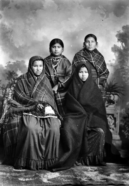 Studio portrait of (sitting, l to r), Lucy Dot Redbird aka Mrs. Jesse John A. Brown (Wak Ja He Win Kah) (Spirit Woman) and Emma Bigbear, Smith-Beaver, Holt (Wau Kon Chaw Zee Win Kaw) (Yellow Thunder Woman) aka Emma Bigbear Holt. They are wrapped in fringed shawls and blankets only showing their faces. Behind them (standing on the right) Jennie Bighawk, Decorra, Manly-Mallory aka Mrs. Edward Manly-Mallory and an unknown woman. They have shawls wrapped around their shoulders and wearing earrings. The woman standing on the left is wearing several necklaces. In the background is a painted backdrop.
