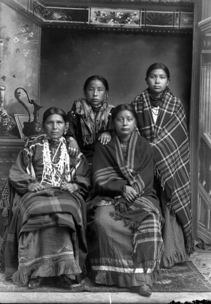 Studio portrait in front of a painted backdrop of two Ho-Chunk women sitting in front of two Ho-Chunk women standing behind them. The woman sitting on the left is wearing several necklaces and earrings, with a shawl on her lap, and the woman sitting on the right is wrapped in a fringed shawl. The woman standing on the left is wearing several necklaces and earrings and short shawl over her shoulders, and the woman on the right is wrapped in a plaid fringed shawl.