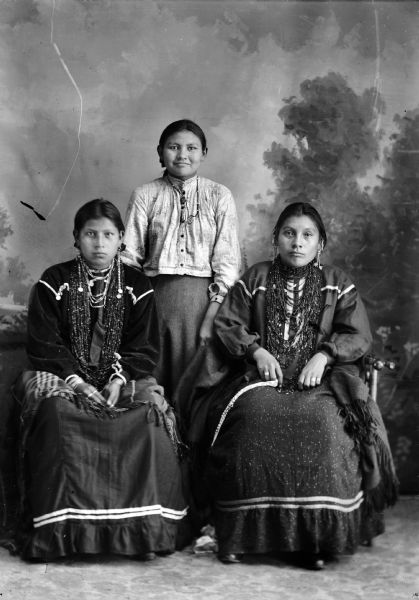 Studio portrait in front of a painted backdrop of three Ho-Chunk women. The two women in front are sitting and wearing traditional dark-colored blouses, several necklaces, earrings, rings, and file bracelets, with shawls on their laps. Behind them in the center a Ho-Chunk woman is standing and wearing a necklace, light-colored blouse and dark-colored skirt.
