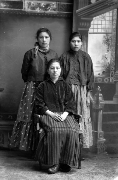 Studio portrait in front of a painted backdrop of a Ho-Chunk woman posing sitting in front of two women standing. The woman sitting has her hands clasped on her lap, and is wearing a dark-colored blouse and striped skirt. The woman standing on the left has her hands behind her back and is wearing a bandana and polka-dot skirt. The woman standing on the right is wearing a skirt and short dark blouse.