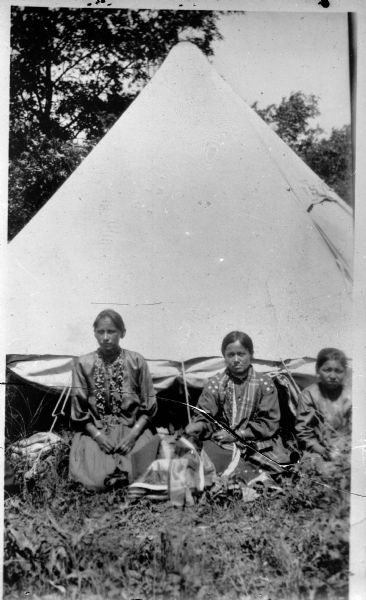 Copy photograph of three young Ho-Chunk women wearing several necklaces posing sitting on the ground outdoors in front of a white tent in a field.