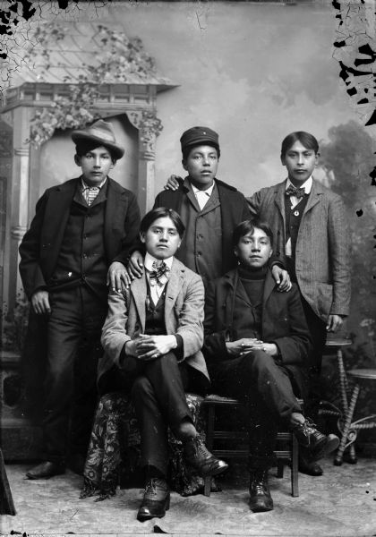 Studio portrait in front of a painted backdrop of (l to r) Johnie Thunder and George Bearchief (Ha Na Kah) sitting with their legs crossed in front of three standing young men. All of the boys are wearing suit coats, and two of the boys are wearing hats.