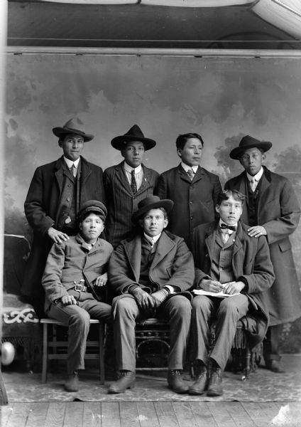 Studio portrait in front of a painted backdrop of three young Ho-Chunk men posing sitting in front of four young Ho-Chunk men posing standing. Most of the men are wearing suit coats and long coats, neckties, and hats, except the man sitting on the far right who is wearing a bow tie.