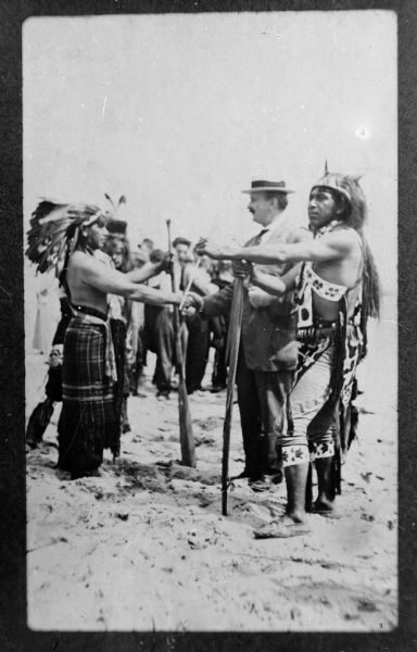 Copy photograph of a European American man shaking hands with a Ho-Chunk man, and several other Ho-Chunk men and European American men standing on sand-covered ground. The European American man shaking hands is probably Art Werner. The two Ho-Chunk men in the front are bare-chested and are wearing regalia such as headdresses and bandoleers.