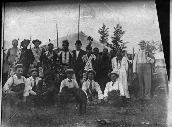 Copy photograph of a large group of people, including several European American men posing sitting, and several Ho-Chunk men and women posing standing behind them. The Ho-Chunk men are dressed in regalia and the European American men are wearing suits and hats. There are also two dogs in front of the group. In the background is a tent.