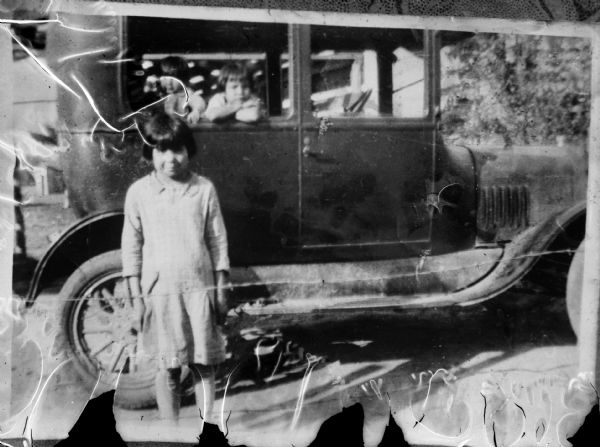 Copy photograph of a Ho-Chunk girl posing standing in front of an automobile, with two Ho-Chunk children peering out the back window of the automobile.