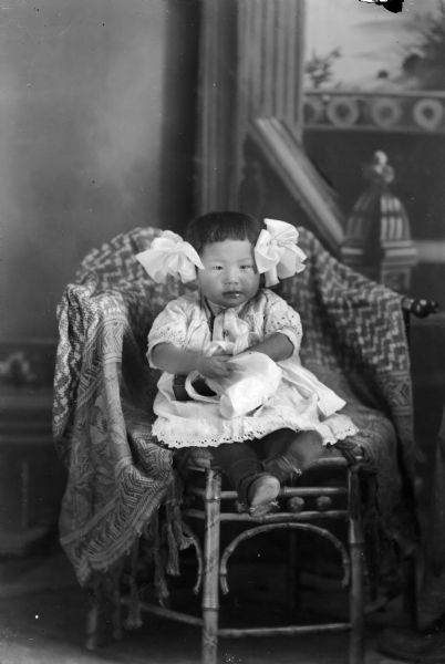 Studio portrait in front of a painted backdrop of a small Ho-Chunk girl posing sitting on a chair draped with a fringed throw. She is wearing a white dress, and two large white ribbons in her hair. She is holding a bonnet in her hands.