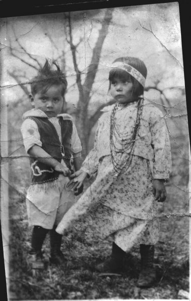 Copy photograph of a Ho-Chunk boy and girl standing in a field. The boy on the left is wearing a ribbon work vest, and the girl is wearing a dress, several necklaces and a beaded head band.