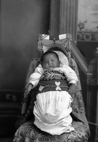 Studio portrait in front of a painted backdrop of a Ho-Chunk infant in a cradle board with ribbon, beadwork, and bell decoration. The cradle board is leaning against the back of a chair. Possibly the child of Adam Thundercloud and Sara Windblowe.
