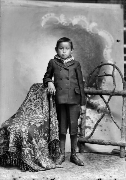 Studio portrait in front of a painted backdrop of a Ho-Chunk boy posing standing in front of a prop wooden fence. He is wearing a winter coat, scarf, and knickers, and has his right arm resting on a chair draped with a fringe throw.