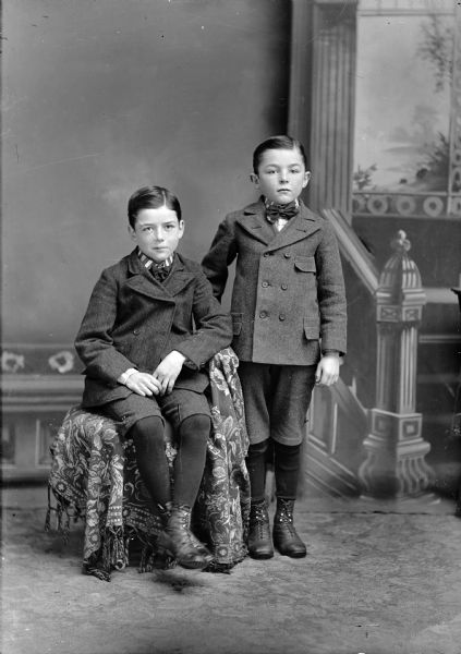 Studio portrait in front of a painted backdrop of two boys wearing winter coats and knickers. The boy on the left is sitting on a chair draped with a fringed throw, and the boy on the right is posing standing.