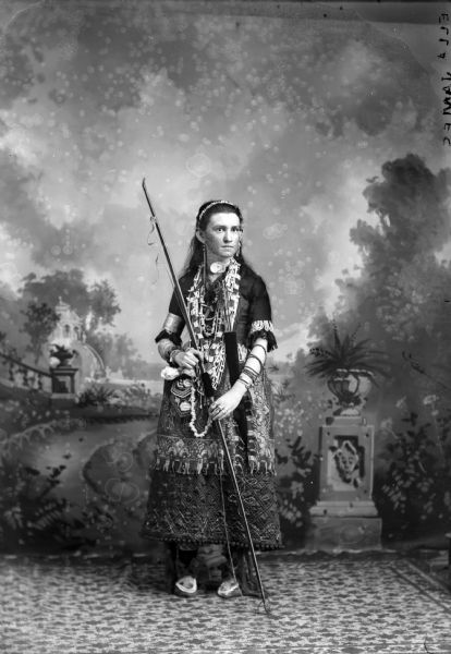 Studio portrait in front of a painted backdrop of a young European American woman posing standing holding an unstrung bow. She is wearing several Ho-Chunk or Sioux objects, including several necklaces, bracelets, earrings, and rings. She is wearing a blouse and a skirt. Identified as Ella James, wearing objects from the collection of Tom Roddy. Negative inscribed in the upper right corner, "Ella James."