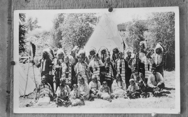 Copy photograph of a large group of Native American/Sioux men, women, and children all wearing regalia, posed outdoors standing and sitting in front of a tipi. The photograph is inscribed in the right corner "Bell Photo."