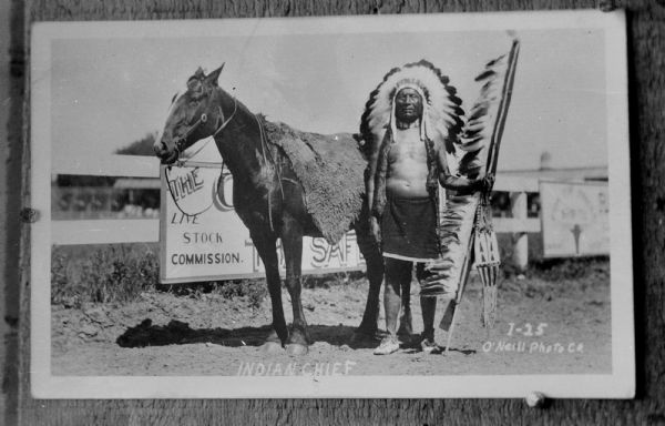 Copy photograph of a Native American/Sioux man posed standing outdoors, holding a feathered spear and wearing a large bonnet. He is standing next to a horse in front of a large sign posted on a fence. The photograph is inscribed in the right corner with : "I-25 O'Neill Photo Co." and in the bottom center, "Indian Chief".