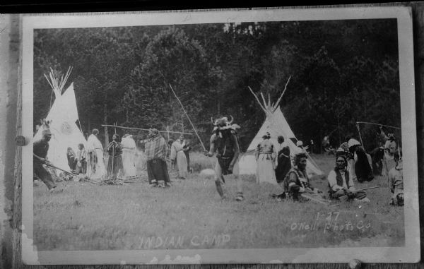Copy photograph of a Native American/Sioux camp and dance ritual, including a man wearing a buffalo horn headdress. In the background there are people sitting and standing near tipis.  The photograph is also inscribed with "Indian Camp, I-27 O'Neill Photo Co."