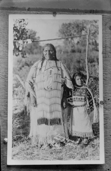 Copy photograph of a Native American/Sioux woman posed outdoors standing with a Native American/Sioux girl. Both are dressed in regalia. The photograph is inscribed "Sioux Squaw and daughter, Bell Photo (c) 148."