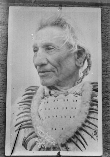 Copy photograph of a Native American/Sioux man with white hair. He is wearing a bear claw necklace. The photograph is inscribed, "Sioux Indian, Black Hills, S. D. Bell Photo (c) 150."