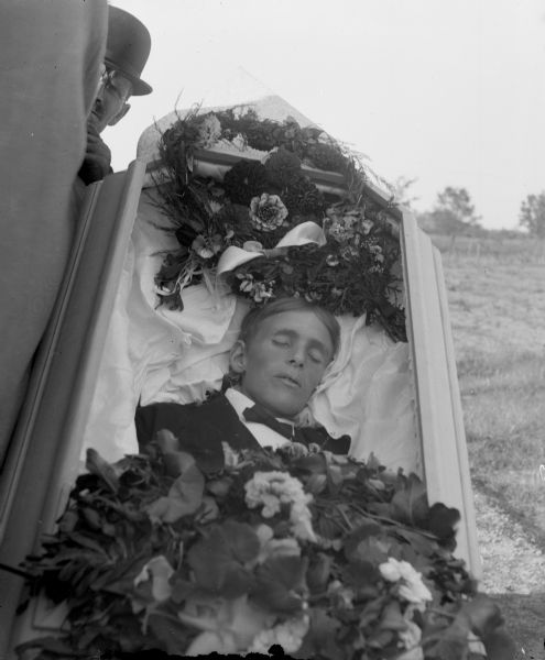 Open casket with the corpse of an European American man lying inside it wearing a dark-colored suit coat and bow tie. Floral arrangements are placed above his head and on top of his midsection. Another man wearing a derby is visible looking from behind the casket.