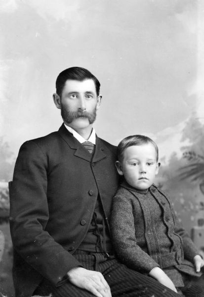 Studio portrait in front of a painted backdrop of a European American man with a large moustache sitting with a young boy leaning against him. The man wears a dark-colored suit coat, vest, striped trousers, and striped necktie. The boy wears a button-down jacket and striped trousers. The man is identified as probably J.J. McGillivray with his son, William McGillivray.