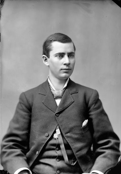 Waist-up studio portrait of a European American man posed sitting wearing a dark-colored suit coat, vest, wide bow tie, and a ribbon watch fob with a pendant in the shape of an Iron Cross. Man identified as John Mills.