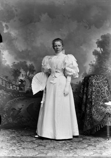 Studio full-length portrait in front of a painted backdrop of a European American woman with light-colored hair posed standing. She is wearing a white or light-colored dress with a ruffled bodice and a high collar, long gloves, holding an open hand fan. Woman identified as Sadie Nolop Dimmick.