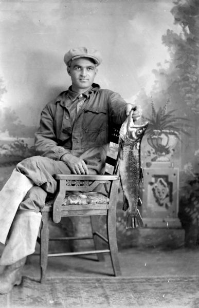 Studio portrait in front of a painted backdrop of a European American man posed sitting sideways in a chair. He is holding a large trout by the mouth with his left hand, and is wearing a light-colored three-quarter length overcoat, trousers, shirt, long boots folded over, and a cap. Identified as probably Royal Ott with a trout from Robinson Creek.