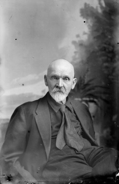 Studio portrait in front of a painted backdrop of an elderly European American man with a gray van dyke beard and bald head posed sitting with his left leg crossed over his left. He is wearing a dark-colored suit coat, shirt, necktie, and trousers. Man identified as probably Ed Wylie.