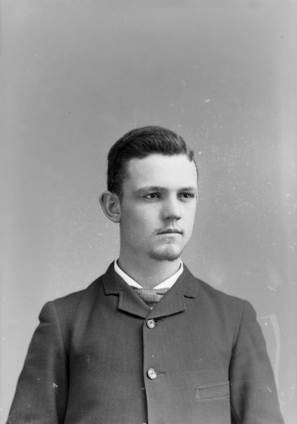 Studio quarter-length portrait of a young European American man with a sparse goatee beard posed wearing a dark-colored, high-buttoned suit coat and checked bow tie. Man identified as Austin Bart.
