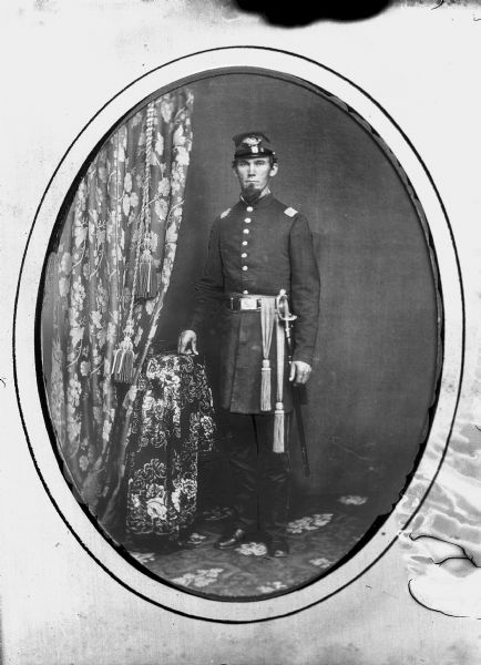Copy photograph of a studio full-length portrait of a European American man posed standing with his right hand resting on a table draped with a floral print cloth near a curtain. He is wearing a military uniform, presumably from the Union army of the United States Civil War, including a forage cap kepi, and a sash and sword in its scabbard. Man identified as probably Captain William Moore.