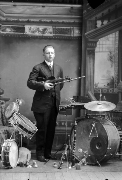 Studio full-length portrait in front of a painted backdrop of a European American man posed standing among various instruments. He is holding a violin under his left arm, and holding a bow in his left hand. He is wearing a dark-colored pattern suit coat, vest, and necktie. Instruments surrounding him include a xylophone, clarinet, bells, and several drums. Man identified as probably Hoften Hagen.
