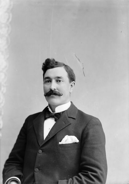 Waist-up studio portrait of a European American man with a handlebar moustache posed sitting. He is wearing a dark-colored suit coat, bow tie, and light-colored handkerchief in his breast pocket. Man identified as Professor J.H. Dersey, who lived in Black River Falls when the High School was built in 1897.